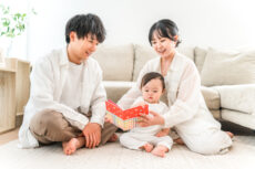 A family sits on a light rug reading a colorful book to their baby on the floor at home.
